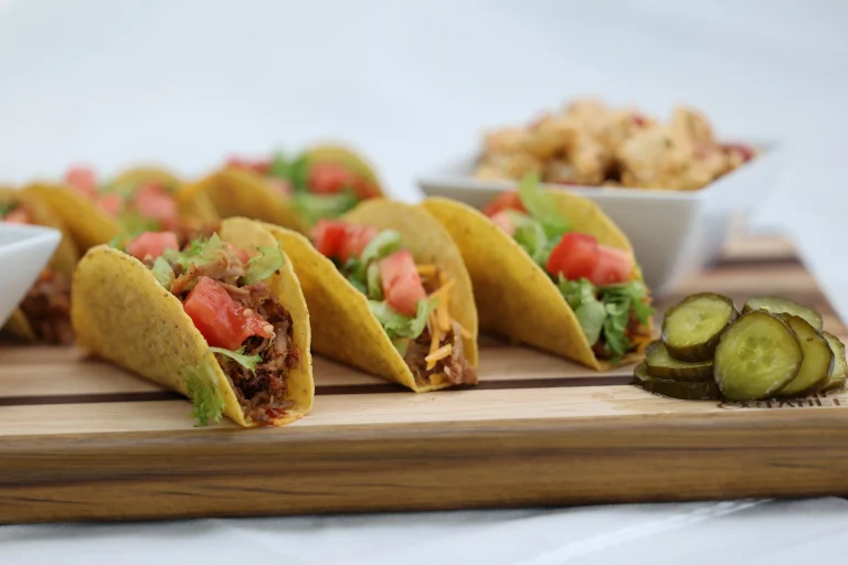 Side view of pulled pork hard-shell tacos on a wooden board with dill pickle slices and side dishes behind them