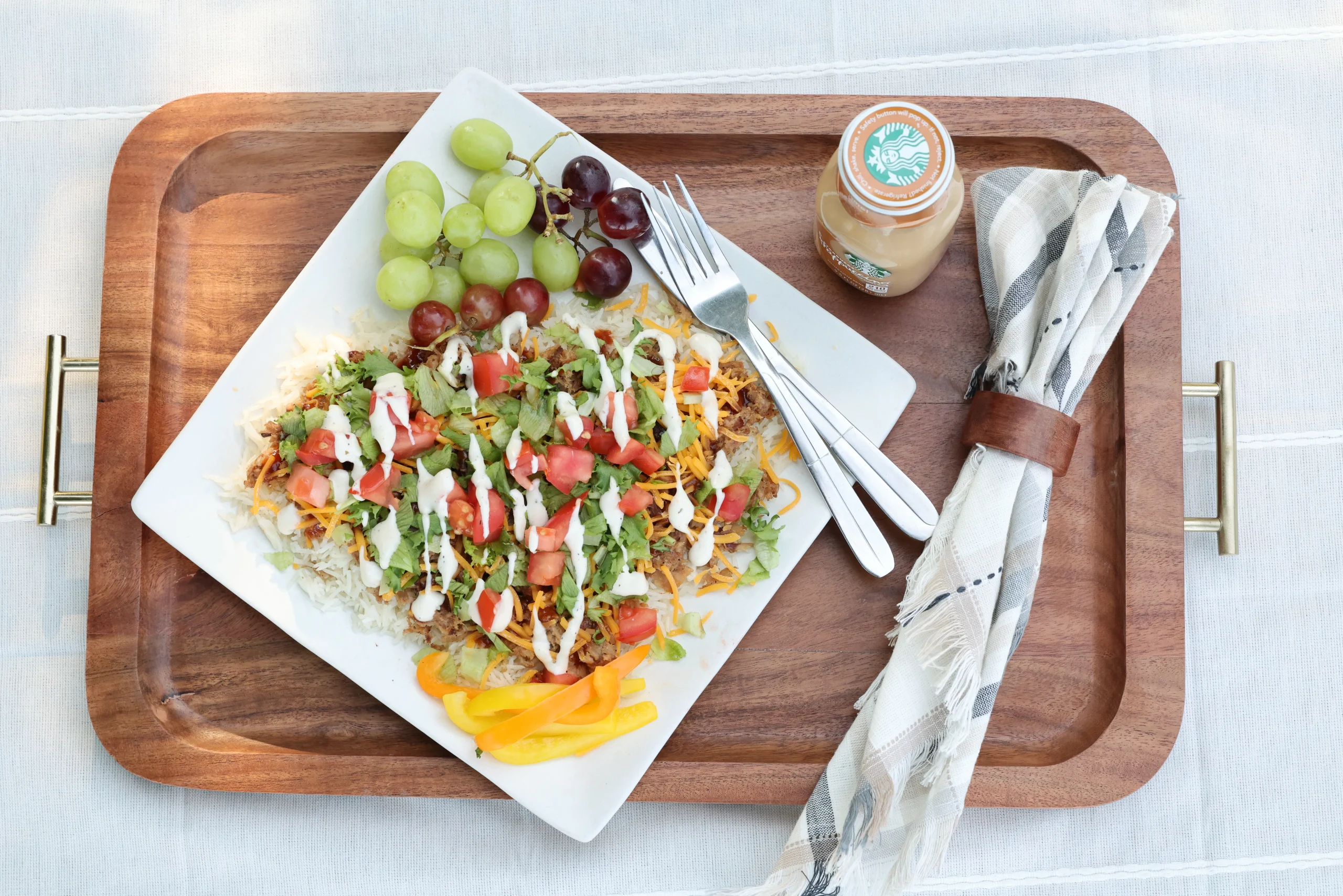 Overhead view of a pulled pork rice bowl topped with shredded cheddar, lettuce, diced tomatoes, and ranch dressing, served on a white plate with grapes and sliced bell peppers, alongside a fork and a small bottle on a wooden serving tray