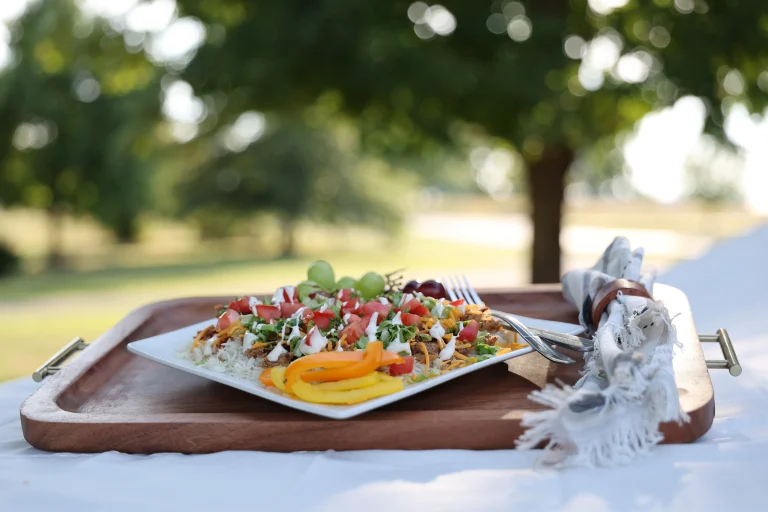Pulled pork rice bowl topped with shredded cheddar, lettuce, diced tomatoes, and ranch dressing, with sliced bell peppers and grapes on the side, plated on a white dish and wooden tray outdoors with green trees in the background